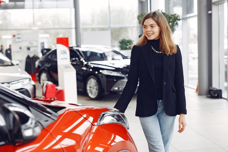 Happy stylish young female in jeans and black jacket standing near modern red shiny car in contemporary car showroom and looking at purchase dreamily