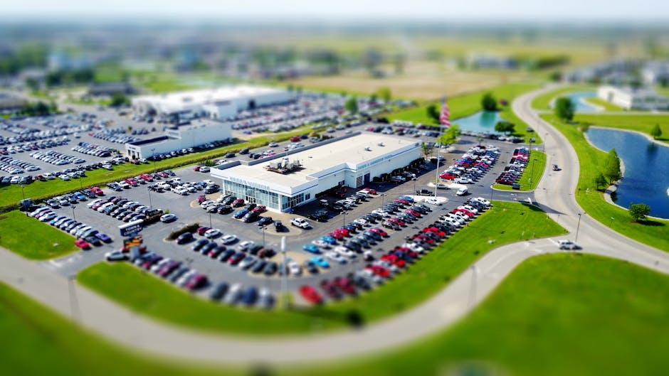 Aerial high-angle view of a bustling car dealership surrounded by parked cars in a green landscape.